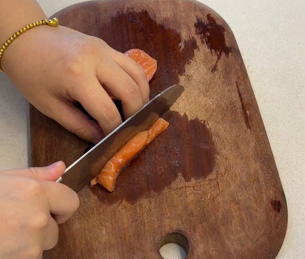 Una mano che utilizza un coltello per tagliare a dadini un filetto di salmone fresco su un tagliere di legno scuro.