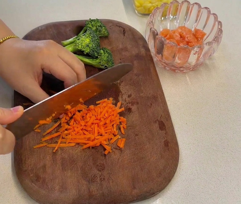 Carote arancioni tritate finemente con un coltello da chef su un tagliere di legno, accanto alle cimette di broccolo preparate in precedenza.