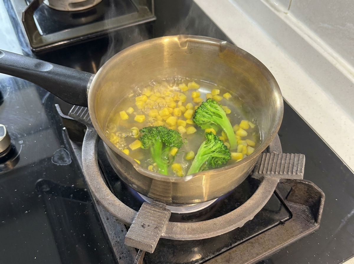 Una pentola in acciaio inossidabile sul fornello piena di acqua bollente, cimette di broccoli e chicchi di mais giallo.