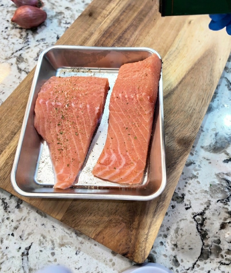 Una mano che condisce i filetti di salmone crudo nella teglia con una spolverata di pepe nero e aglio.