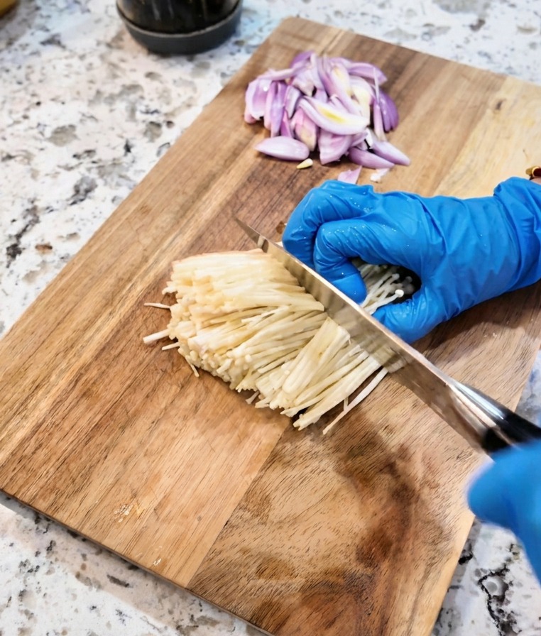 Un coltello taglia la base terrosa di un mazzo di funghi enoki bianchi su un tagliere.
