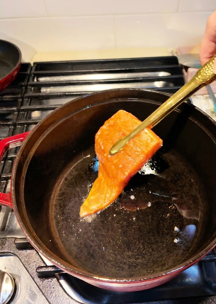 I filetti di salmone marinati vengono adagiati in una pentola di ghisa nera preriscaldata con delle pinze da cucina.