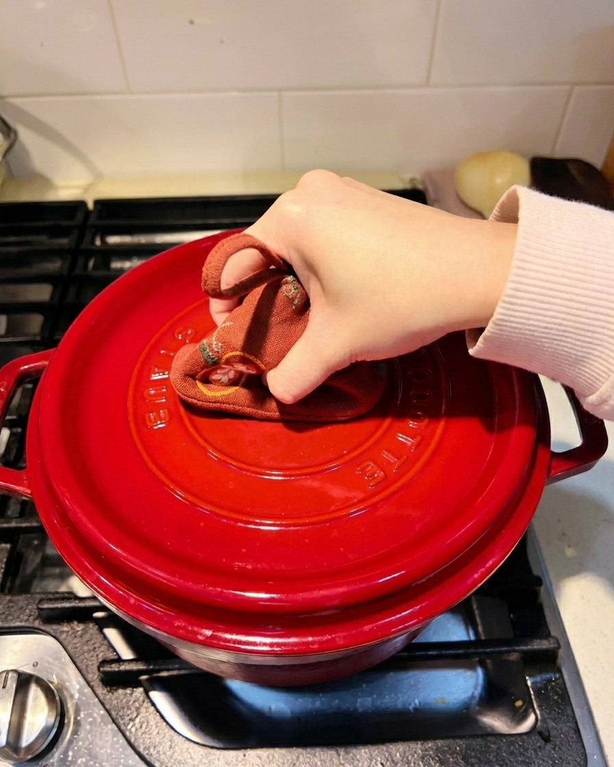 Una mano che usa una presina colorata per chiudere saldamente il coperchio di una pentola in ghisa rossa sul fornello.
