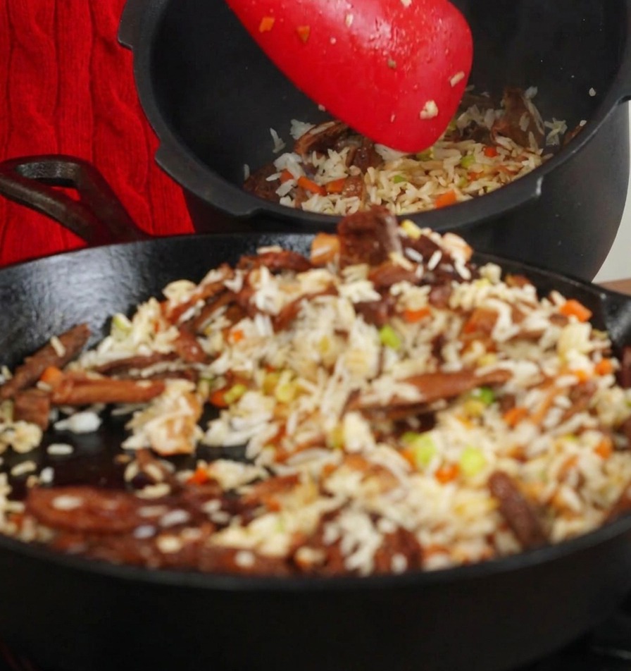 Spatola rossa che trasferisce il riso saltato e i funghi dalla padella al cestello interno di un cuociriso nero.