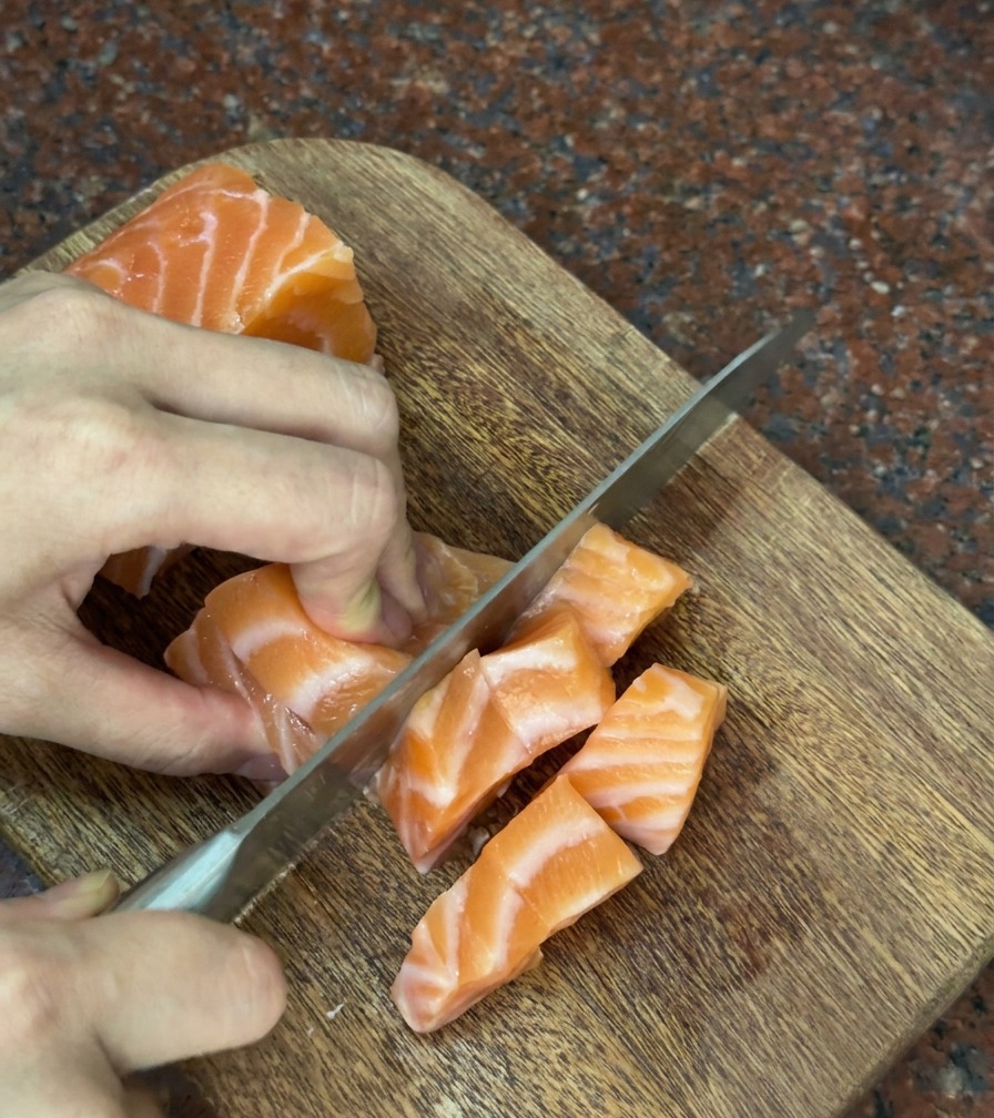 Mani che tagliano con cura un pezzo di salmone fresco in cubetti spessi e regolari su un tagliere di legno.