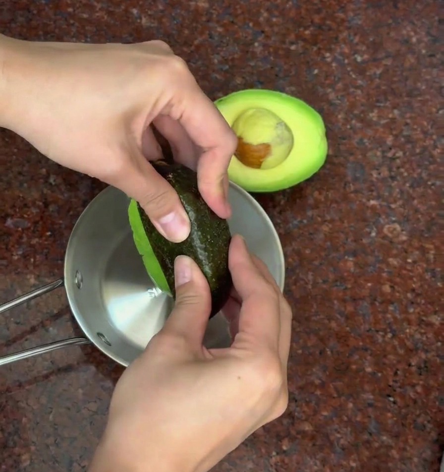 Una persona che rimuove la buccia scura dalla polpa verde di un avocado tagliato a metà sopra una ciotola di metallo.