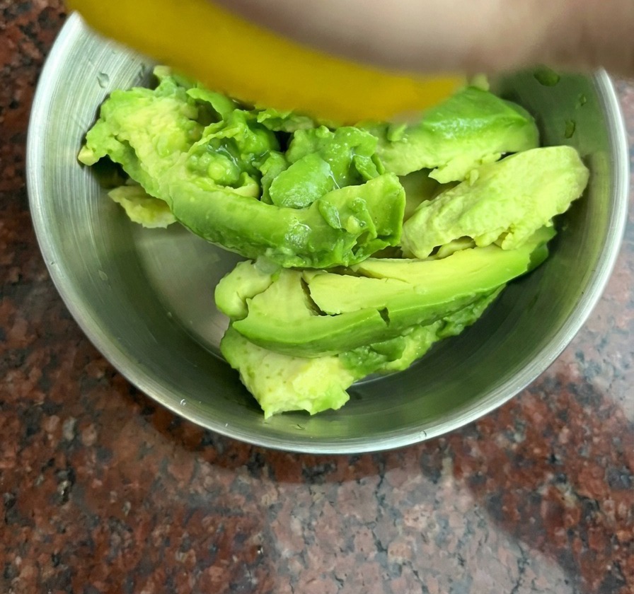 Una mano che spreme un limone giallo sopra pezzi di avocado in una ciotola di acciaio inossidabile.