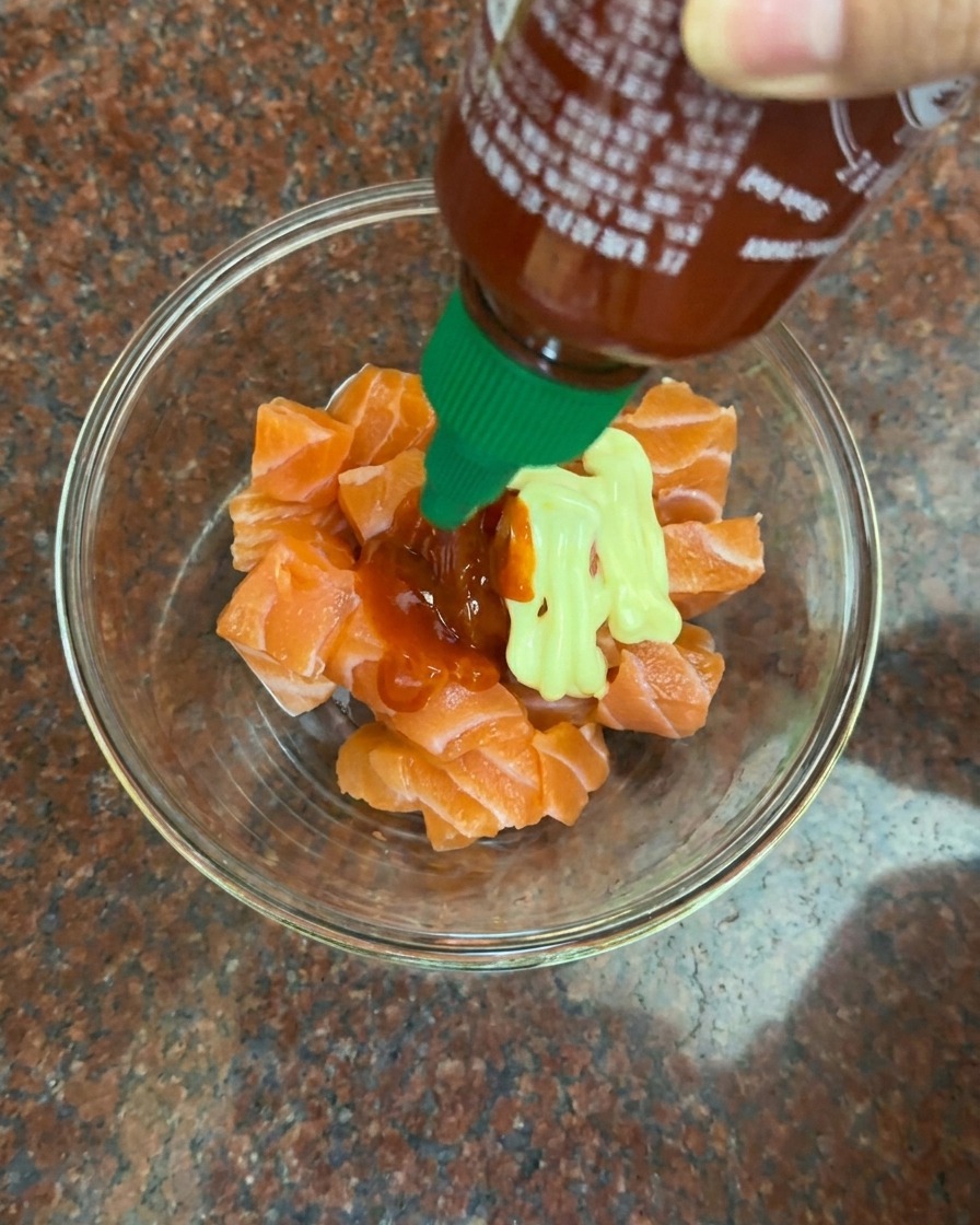 Aggiunta di maionese e salsa chili rossa a una ciotola con cubetti di salmone crudo.