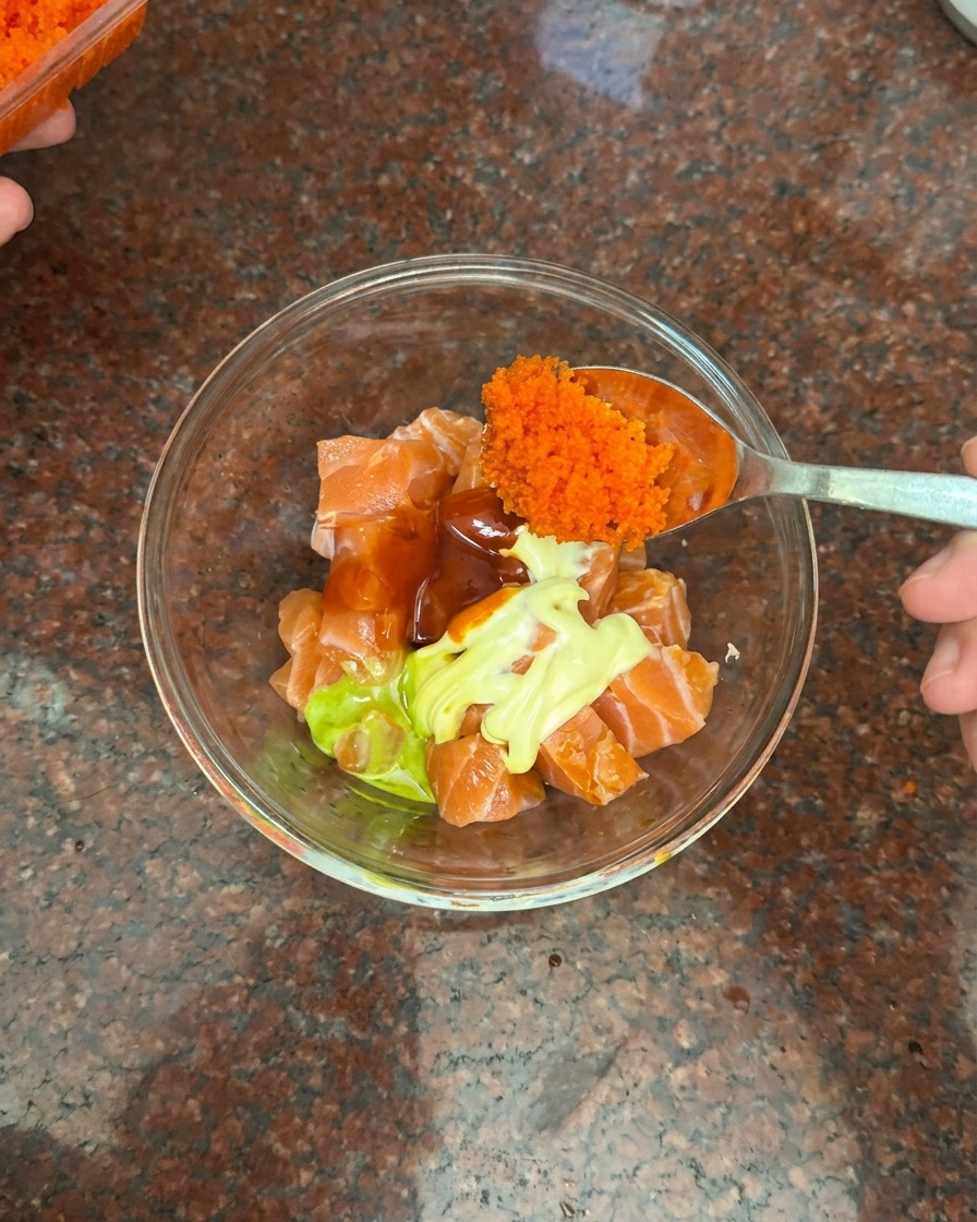 Un cucchiaio che aggiunge uova di pesce arancioni a una ciotola con salmone, maionese e salsa chili.