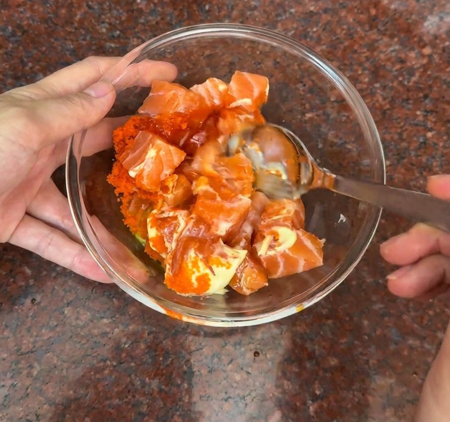 Una ciotola di vetro con cubetti di salmone mescolati a uova di pesce e una salsa cremosa arancione.
