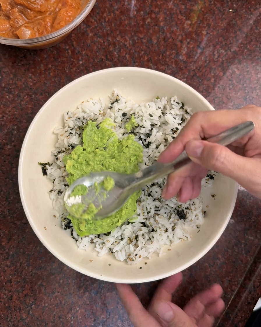 Un cucchiaio che stende crema di avocado verde su un letto di riso bianco e scaglie di alga nori in una ciotola bianca.