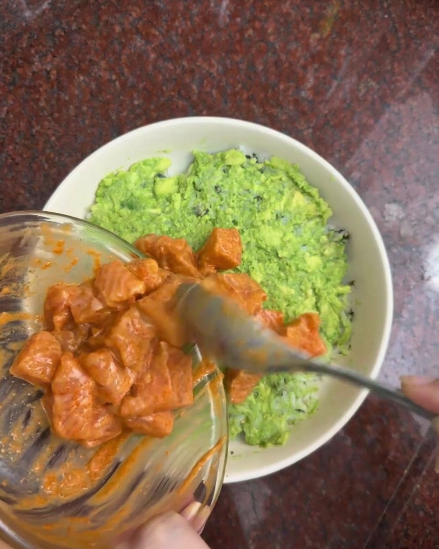 Cubetti di salmone marinati che vengono trasferiti da una ciotola di vetro su uno strato di avocado in una ciotola da portata.