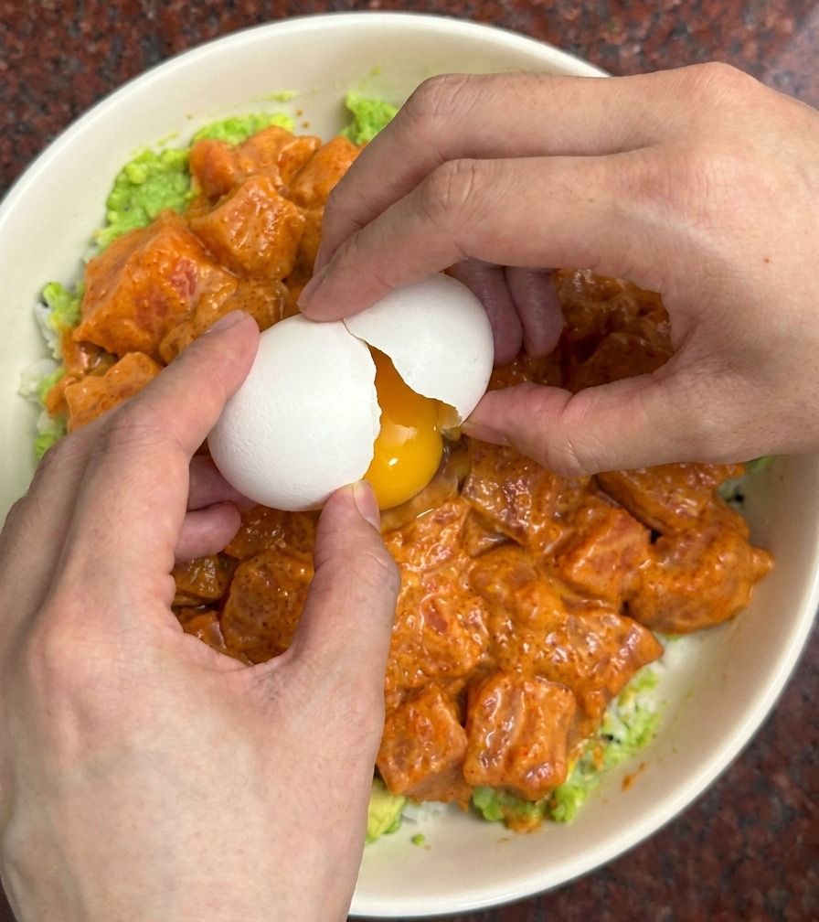 Due mani che rompono un guscio duovo sopra una ciotola colorata con salmone e avocado.