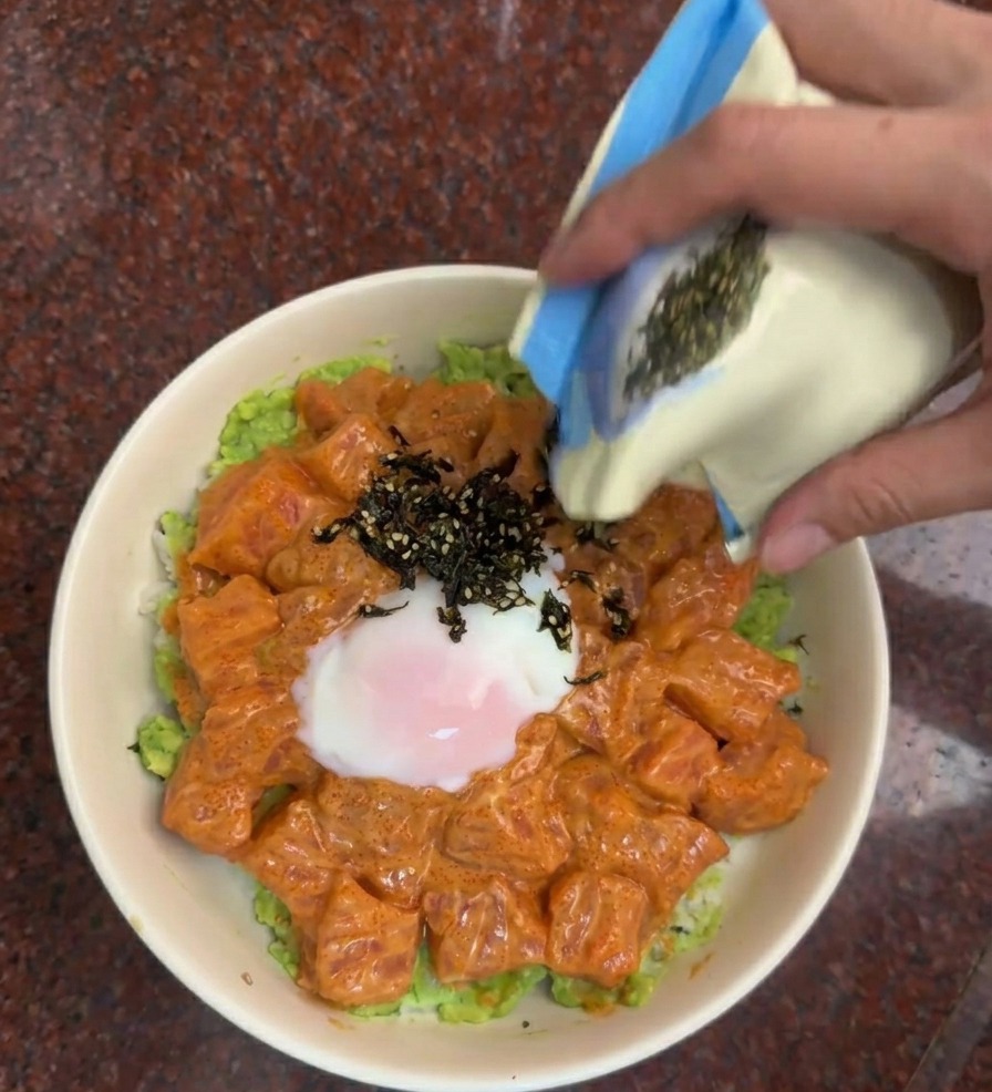 Una mano che versa un mix di strisce di alga nera e semi di sesamo da una bustina sulla bowl di salmone.