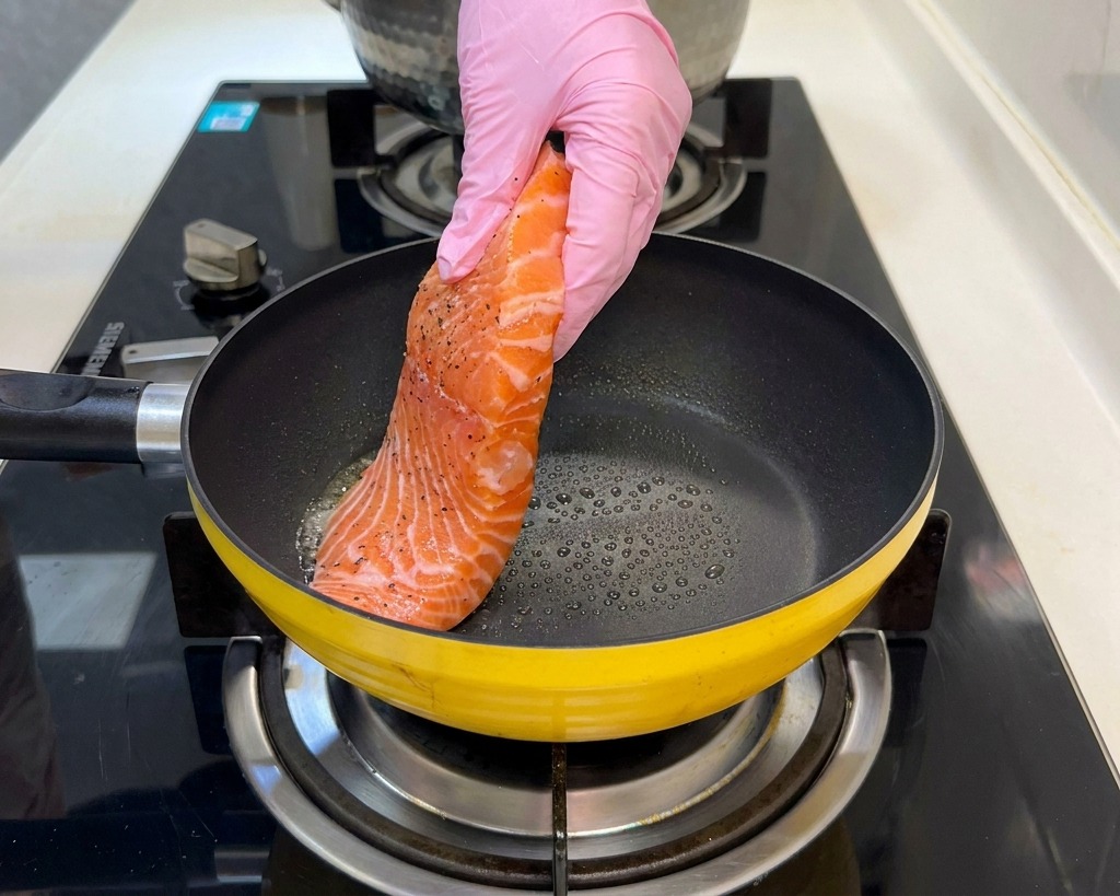 Un filetto di salmone crudo condito viene adagiato in una padella gialla calda da una mano con guanto rosa.