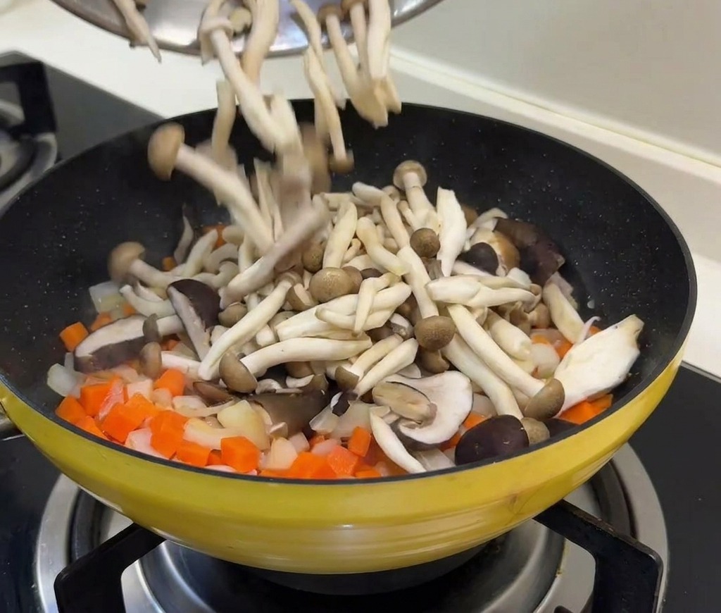 Funghi freschi e piselli versati da una ciotola in una padella con carote e cipolle soffritte.
