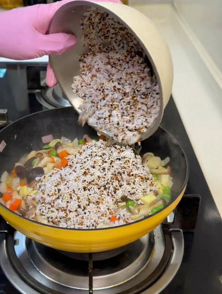 Una mano versa una ciotola di riso bianco e quinoa in una padella con verdure e funghi.