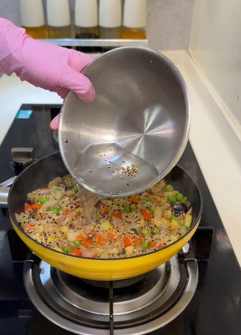 Acqua versata da una ciotola dacciaio in una padella gialla piena di riso e verdure colorate.