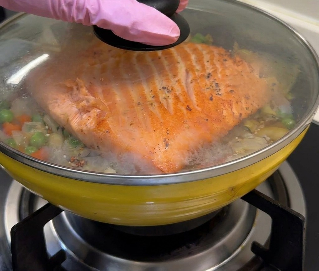 Una padella gialla con salmone e riso viene coperta con un coperchio di vetro trasparente.