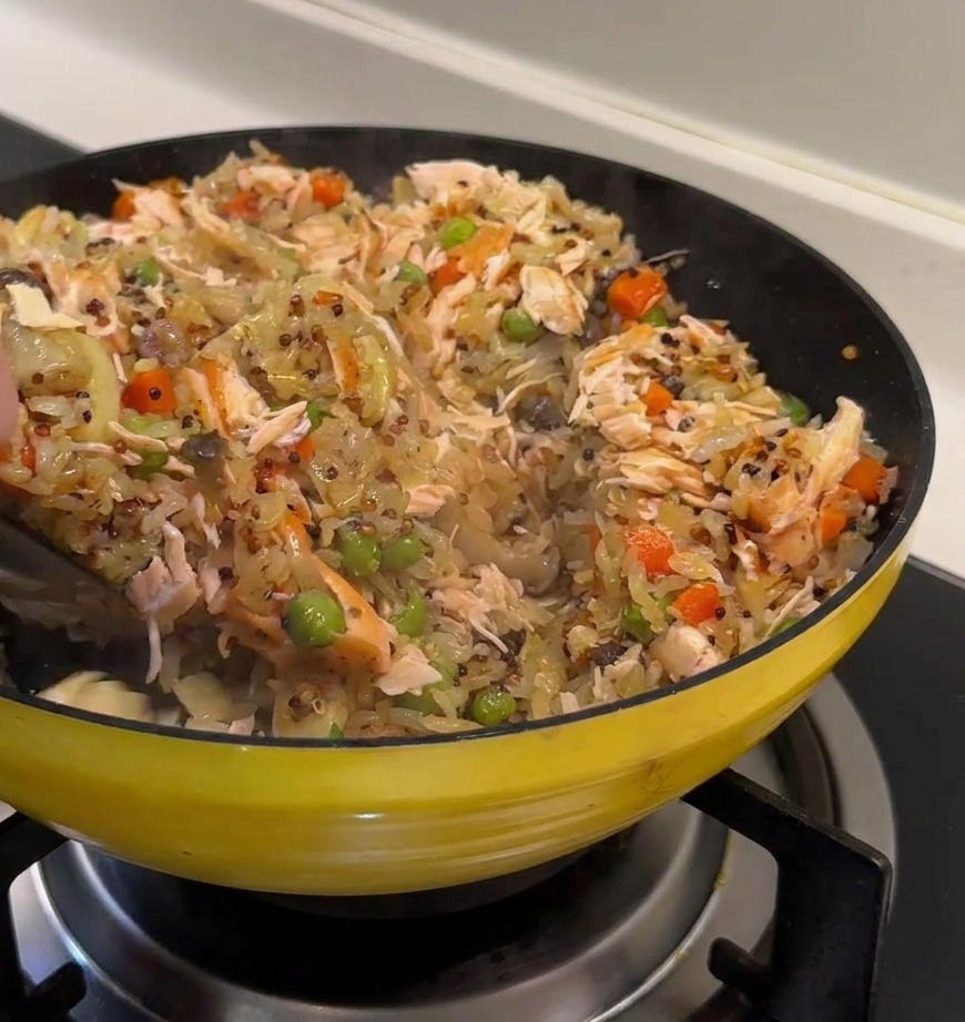 Una spatola nera incorpora grandi pezzi di salmone in una padella colorata con riso, quinoa, piselli e carote.