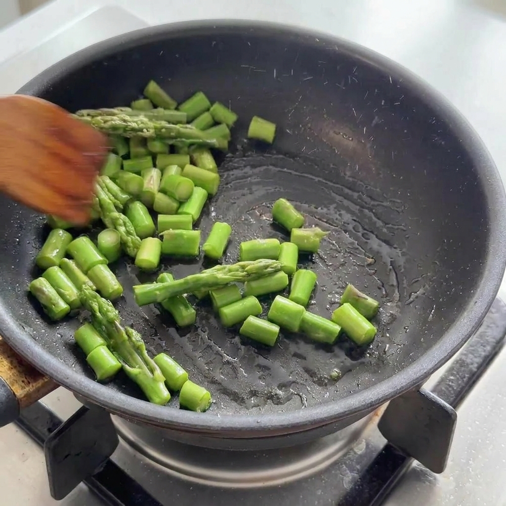 Pezzetti di asparagi verdi saltati in una padella nera con una spatola di legno.