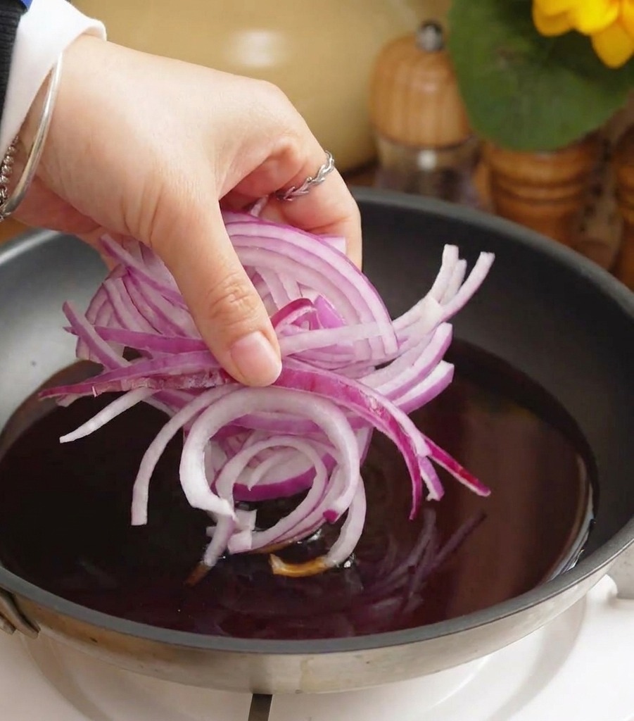 Una mano che versa una manciata di cipolle rosse affettate in una padella piena di salsa scura che sobbolle.