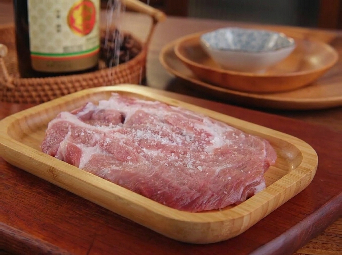Sale fino e pepe nero cosparsi su un pezzo di maiale crudo appoggiato su un vassoio di legno.