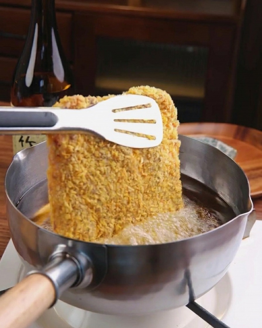Una cotoletta impanata immersa in una pentola di olio bollente usando pinze di metallo.