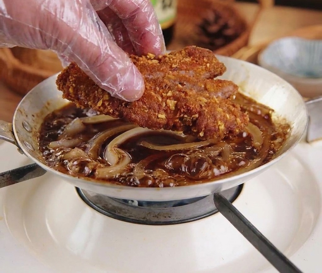 Una mano posiziona la cotoletta affettata sopra le cipolle che sobbollono nella salsa scura.