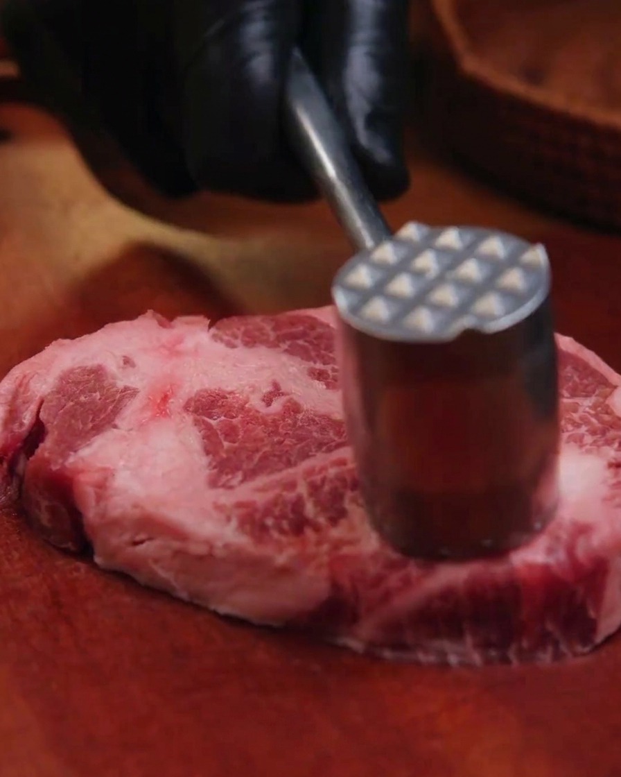 Una mano con guanto nero usa un batticarne in metallo per intenerire una fetta spessa di coppa di maiale cruda su un tagliere di legno.