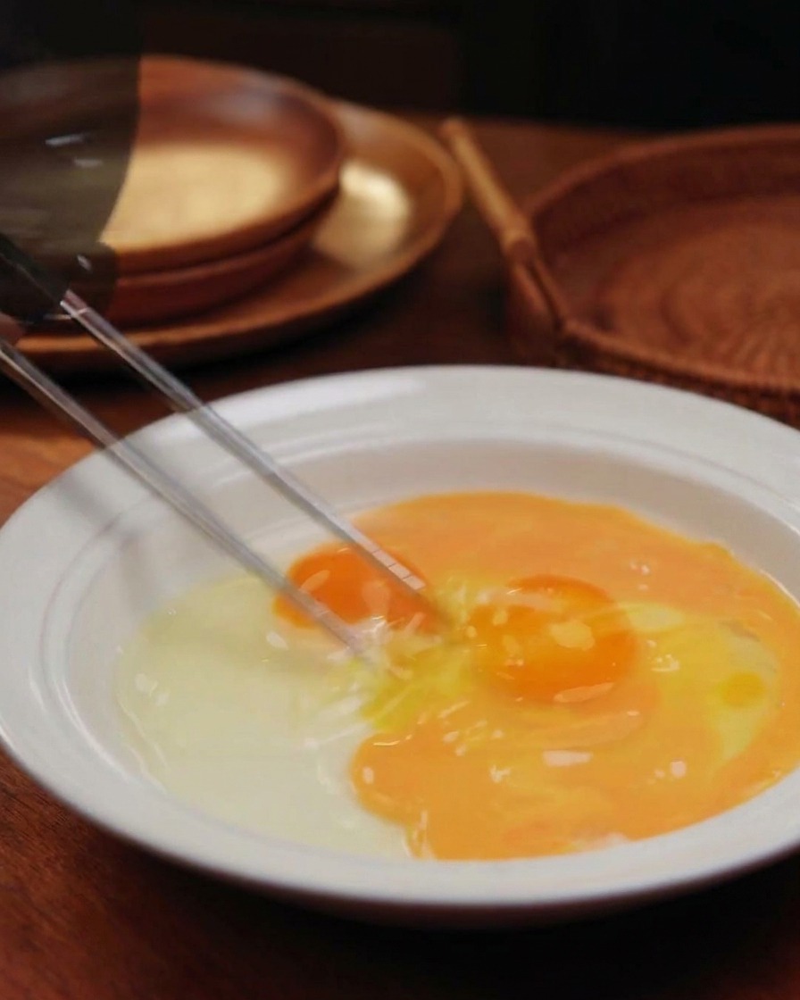 Bacchette che sbattono due uova in una ciotola bianca, creando un composto striato di giallo e arancione.