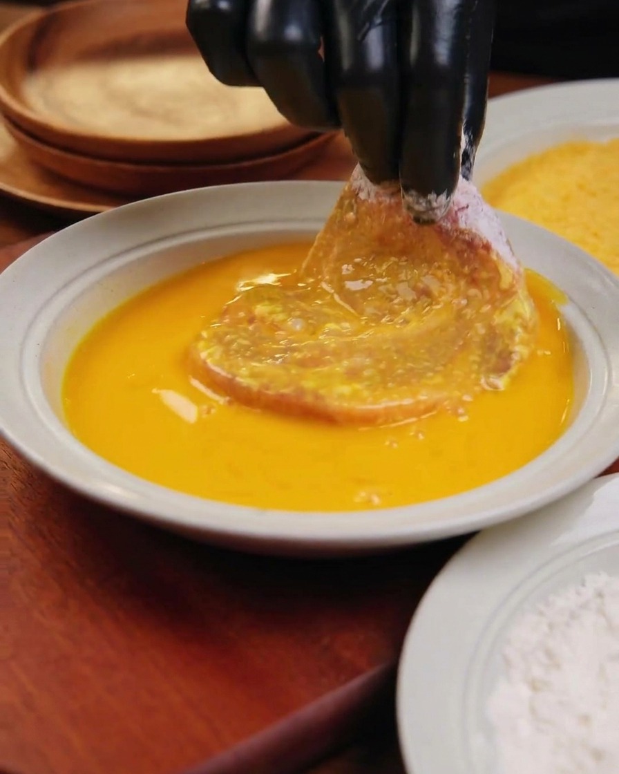 Una mano con guanto nero immerge una grossa cotoletta di maiale infarinata in una ciotola bianca con uovo sbattuto giallo vibrante.