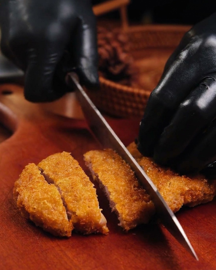Un coltello affilato che taglia una cotoletta di maiale fritta dorata su un tagliere di legno tenuto da mani con guanti neri.