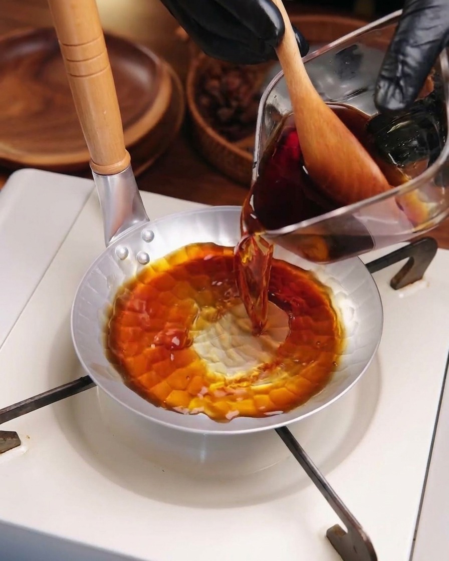 Salsa scura per katsudon versata da un misurino di vetro in una piccola padella di metallo rotonda su un fornello portatile.