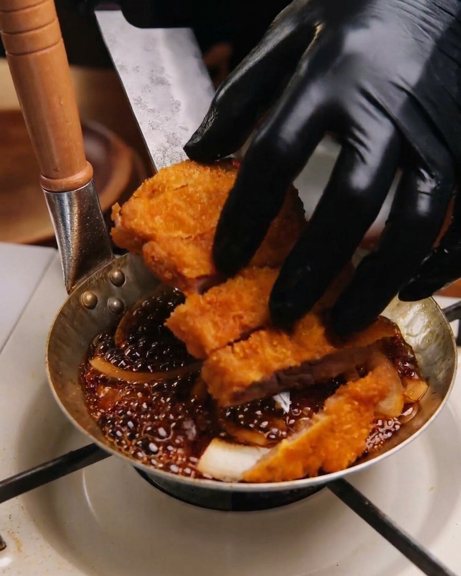 Mani con guanti neri che sistemano i pezzi di cotoletta fritta in una padellina sopra un letto di cipolle e salsa scura.