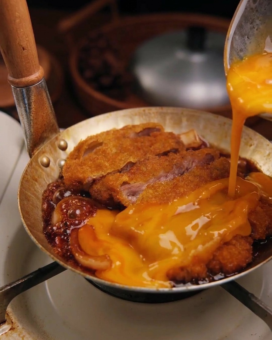 Composto di uova crude appena sbattute versato da una ciotola sopra una cotoletta di maiale che sfrigola nella salsa con le cipolle.