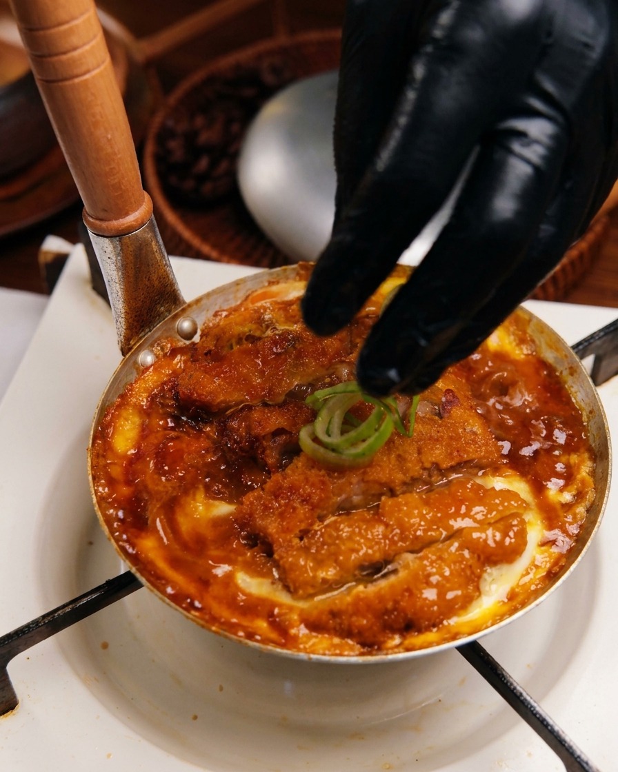 Una mano guantata che posiziona anelli di cipollotto fresco sopra il Katsudon pronto in una piccola padella di metallo.