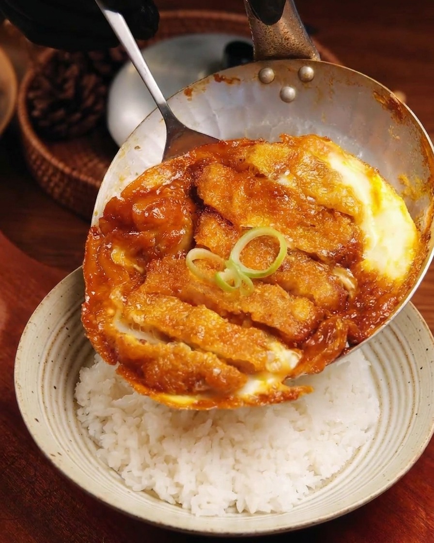 Il composto di cotoletta e uova completato che scivola dalla padella su una ciotola di riso bianco soffice.