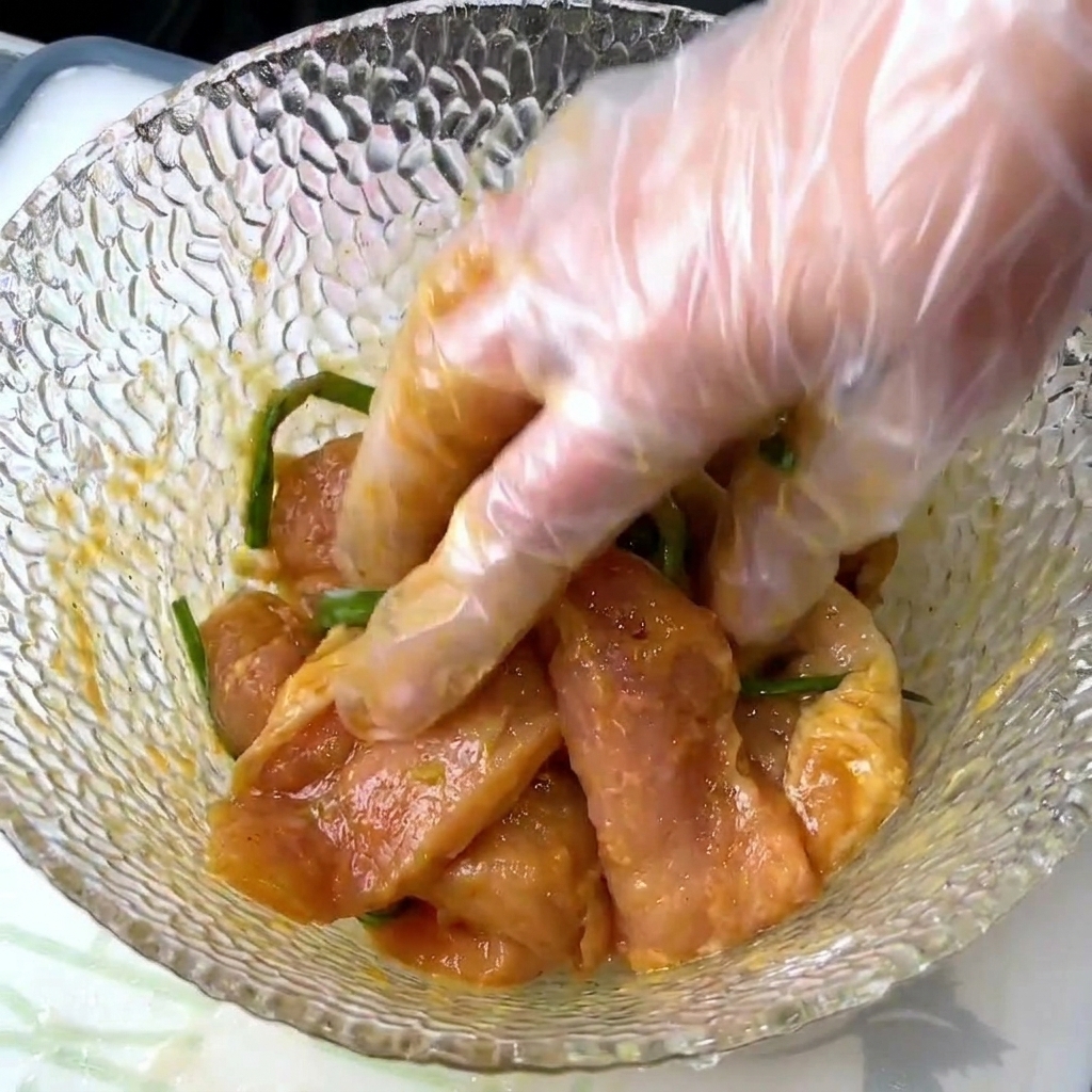 Una mano guantata mescola fette di maiale con cipollotti e condimenti liquidi in una ciotola di vetro.