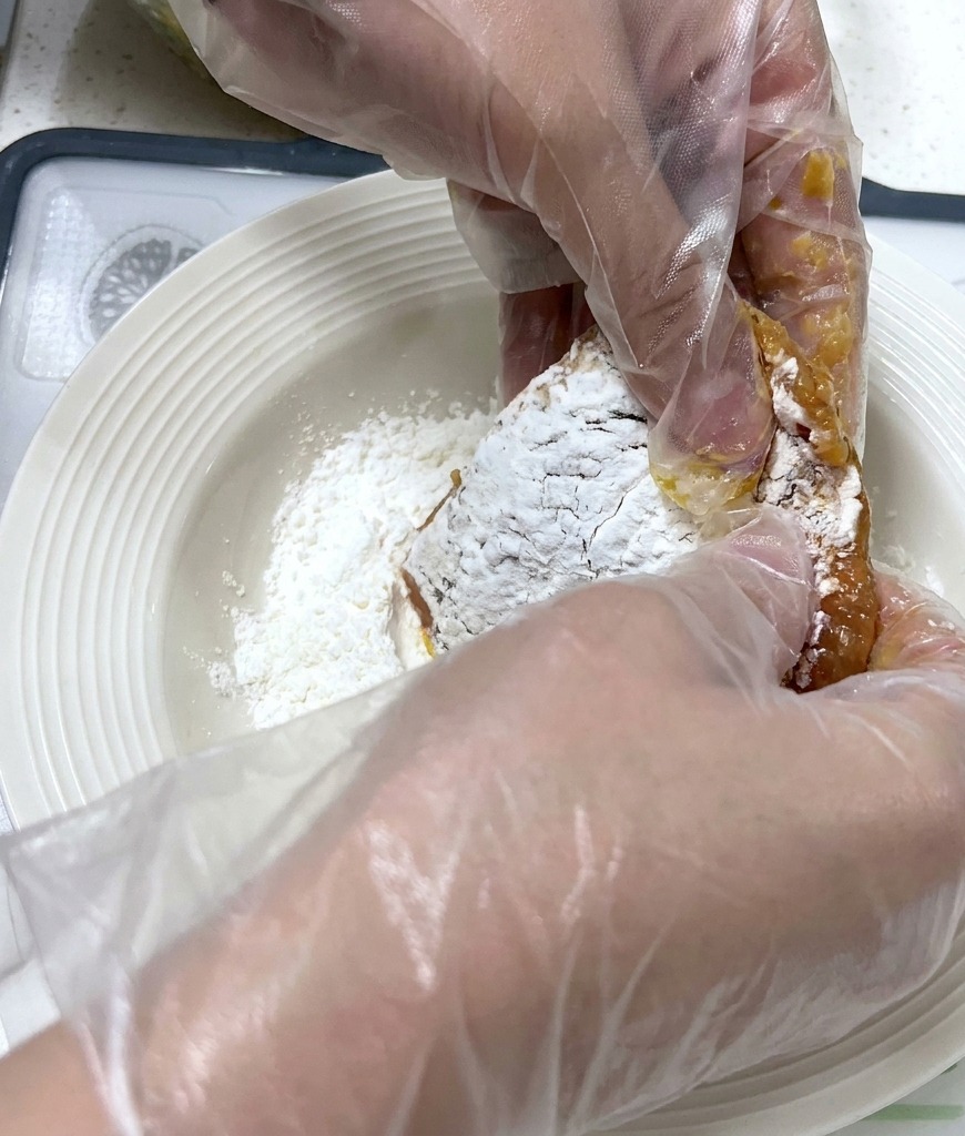 Mani guantate che premono una cotoletta di maiale marinata in un piatto con polvere di amido bianca.