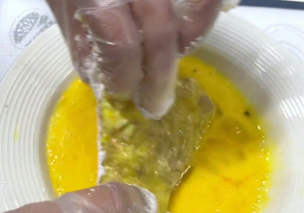 Mani che immergono una cotoletta infarinata in un piatto con uovo sbattuto di colore giallo brillante.