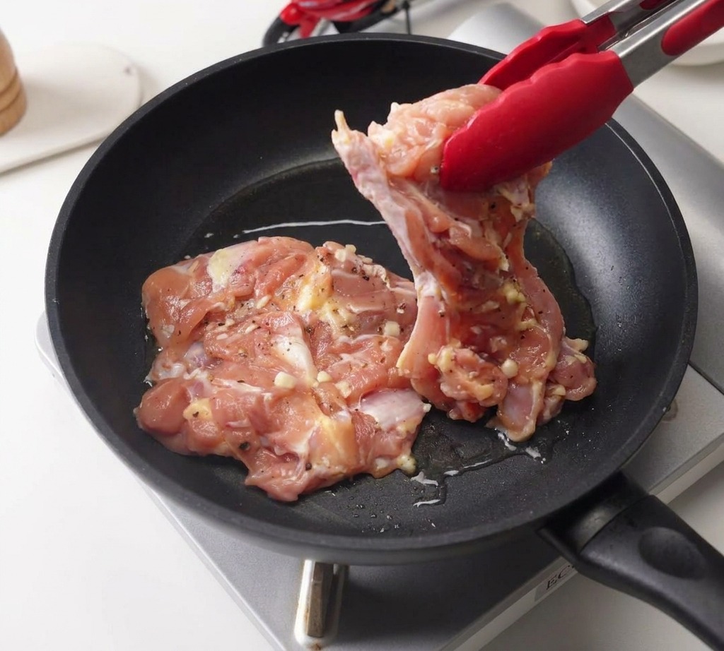 Pinze rosse che adagiano una sovracoscia di pollo marinata in una padella nera con un velo dolio.