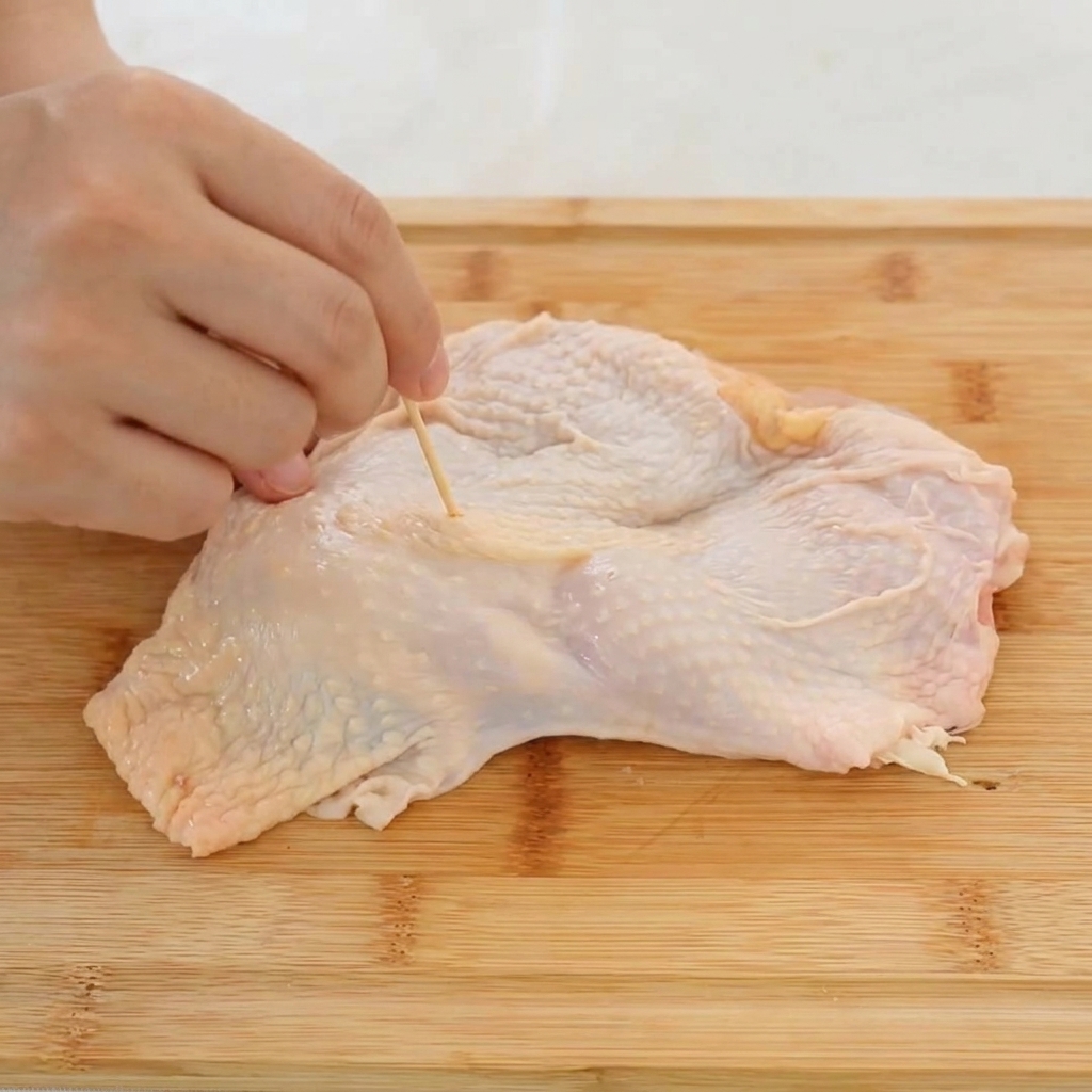 Primo piano di una mano che usa uno stuzzicadenti di legno per praticare fori nella pelle di una sovracoscia di pollo cruda.