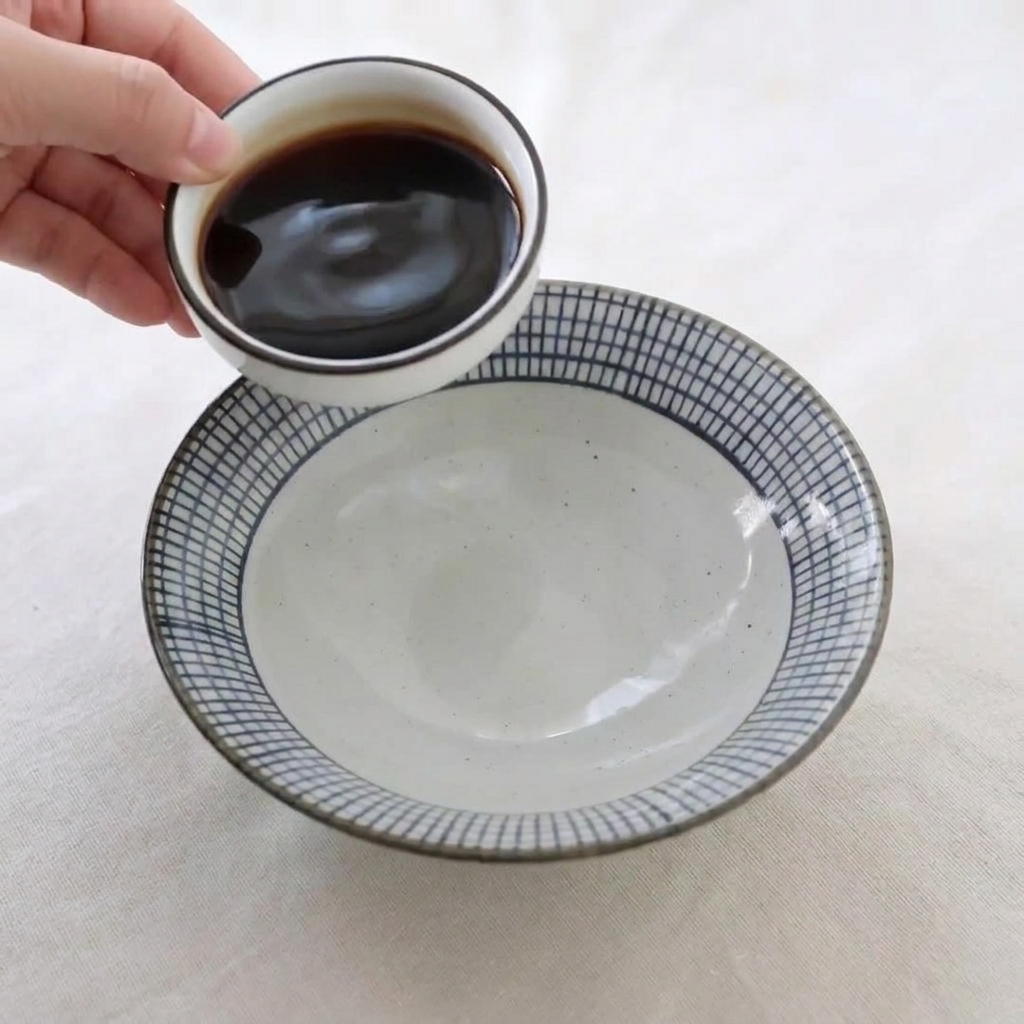 Una mano che versa condimenti liquidi scuri da un piattino bianco in una ciotola di ceramica più grande per preparare la glassa al miele.