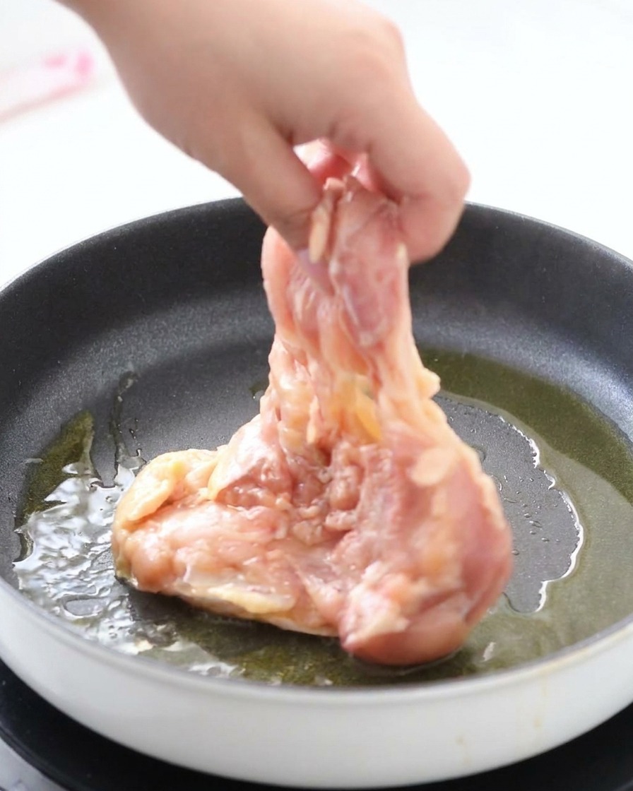 Una mano che adagia una sovracoscia di pollo marinata con la pelle rivolta verso il basso in una padella calda con olio sfrigolante.