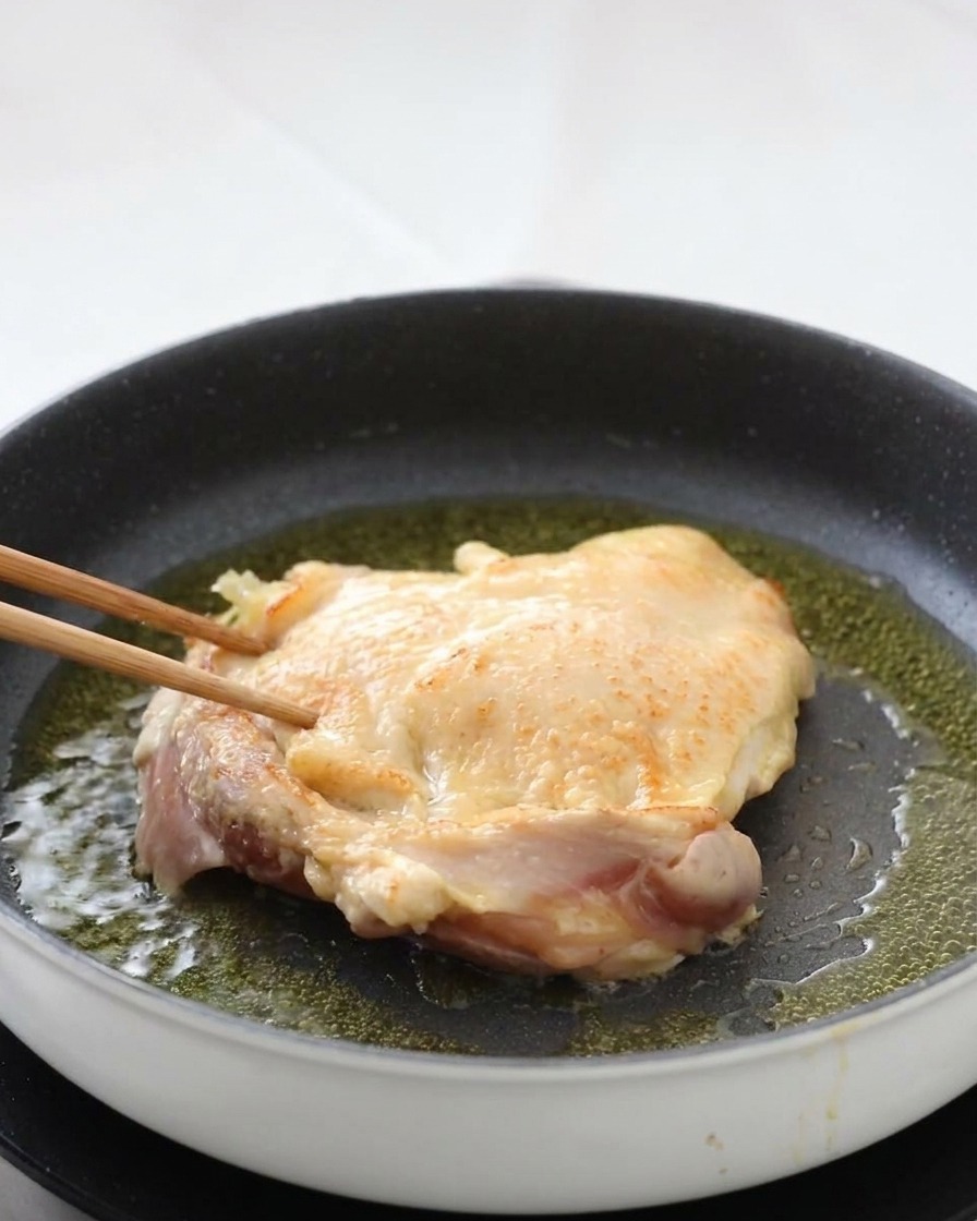 Una sovracoscia di pollo girata in padella con bacchette di legno, rivelando una superficie della pelle perfettamente dorata e croccante.