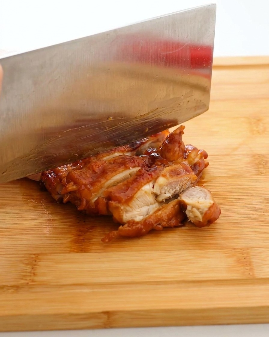 Un grande coltello da cucina che taglia a strisce una sovracoscia di pollo lucida e glassata su un tagliere di legno.