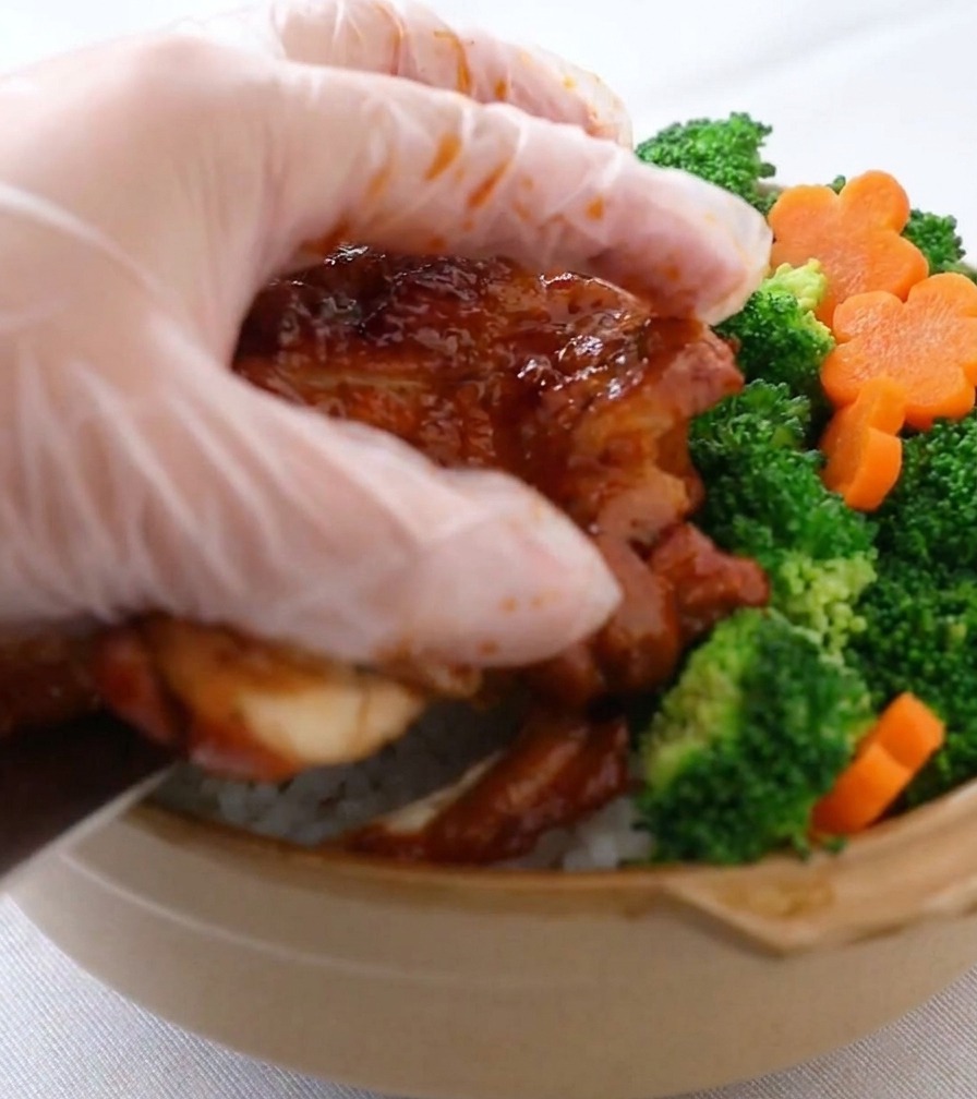Una mano con un guanto di plastica trasparente che posiziona broccoli verde brillante e carote a fiore in una ciotola di ceramica già riempita di riso bianco e strisce di pollo glassato.