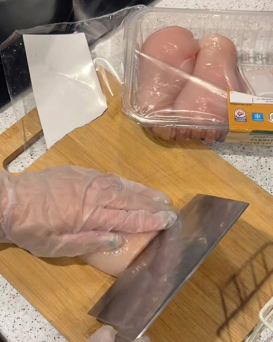 Una mano con un guanto di plastica trasparente affetta un petto di pollo crudo in filetti sottili con una mannaia su un tagliere di bambù.