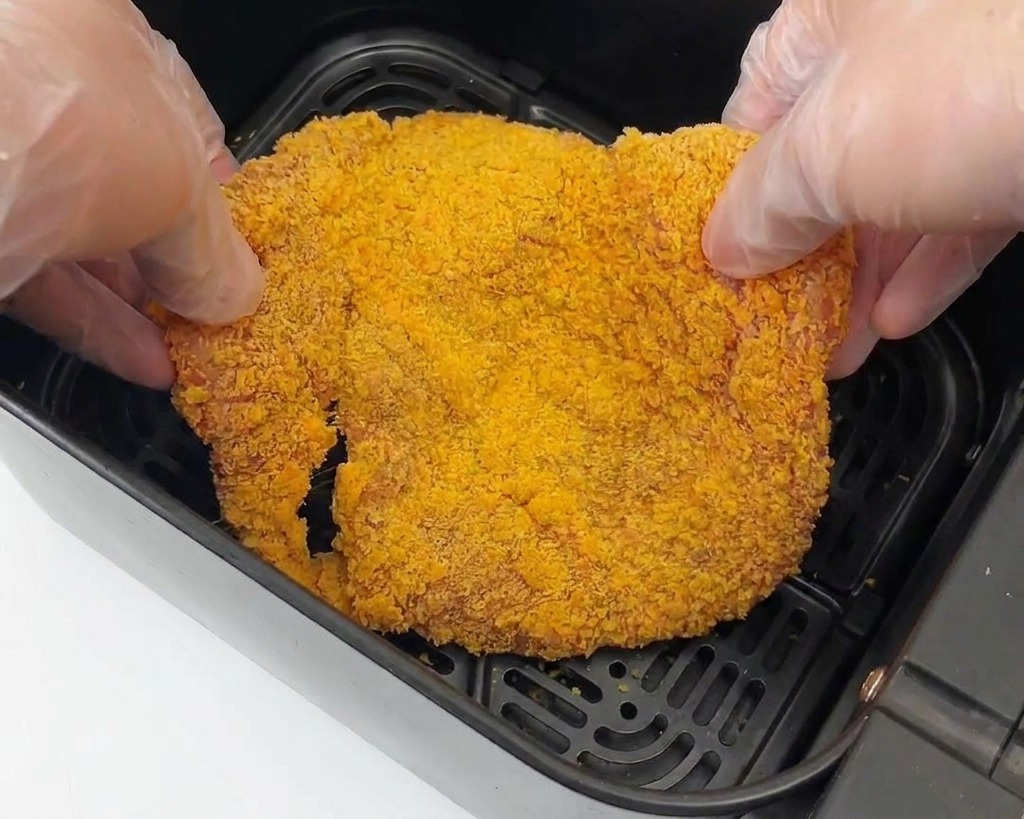 Due mani guantate che posizionano un pezzo di pollo impanato nel cestello nero di una friggitrice ad aria.